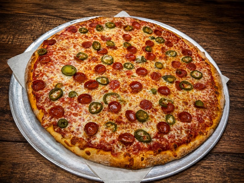 Whole pepperoni and jalapeño pizza on a metal tray, resting on a rustic wooden table.