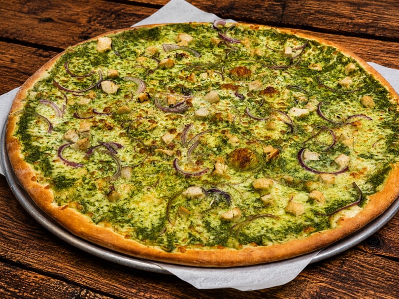 Large pesto chicken pizza with red onions on a wooden table.