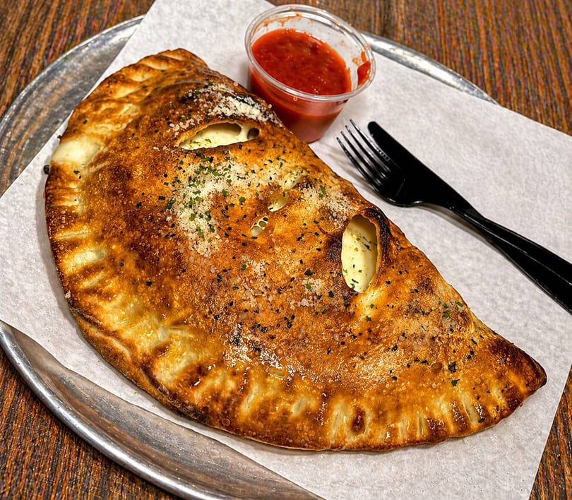 Golden-brown calzone with melted cheese, served with a side of marinara sauce and black cutlery.